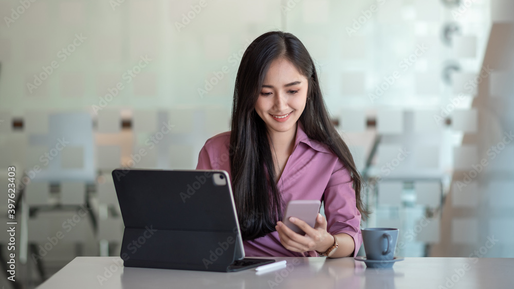 Fototapeta premium Young asian businesswoman beautiful charming smiling and using tablet and mobile phone, while sitting at table and working in office.