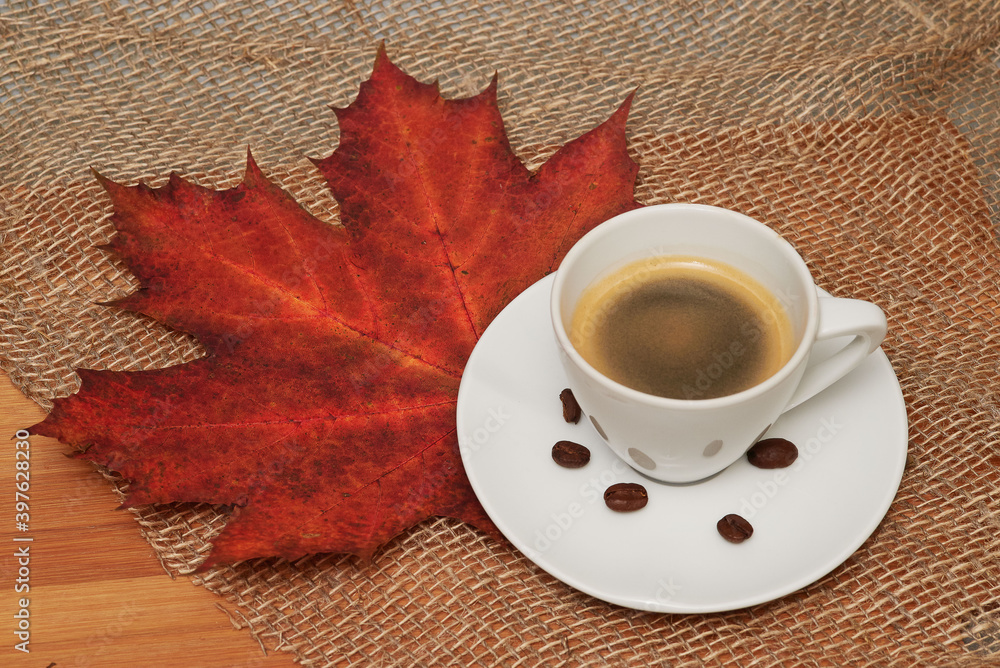 A cup of black coffee and coffee beans with canvas and red leaf as background