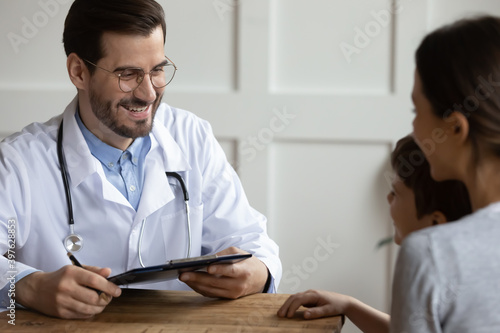 Smiling Caucasian young male doctor or GP talk consult little boy patient at checkup in hospital with mom. Caring man pediatrician speak examine small child at clinic consultation. Healthcare concept.