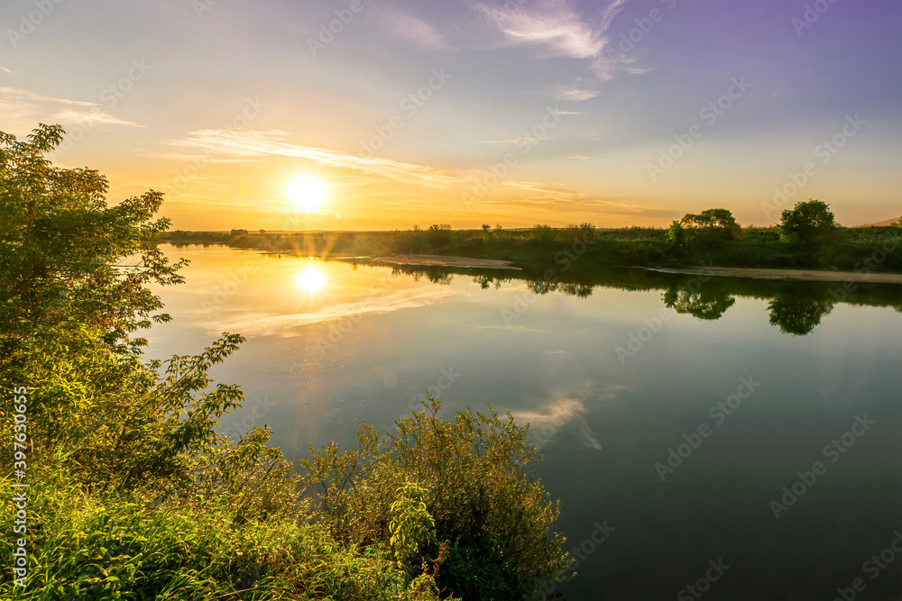 Fototapeta premium Scenic view at beautiful summer river sunset with reflection on water with green bushes, grass, golden sun rays, calm water ,deep blue cloudy sky and glow on a background, spring evening landscape