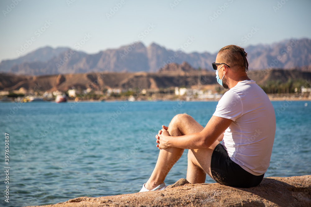 man in a medical mask sits on the beach. tourist man wearing surgical face mask enjoy beautiful sea and mountains