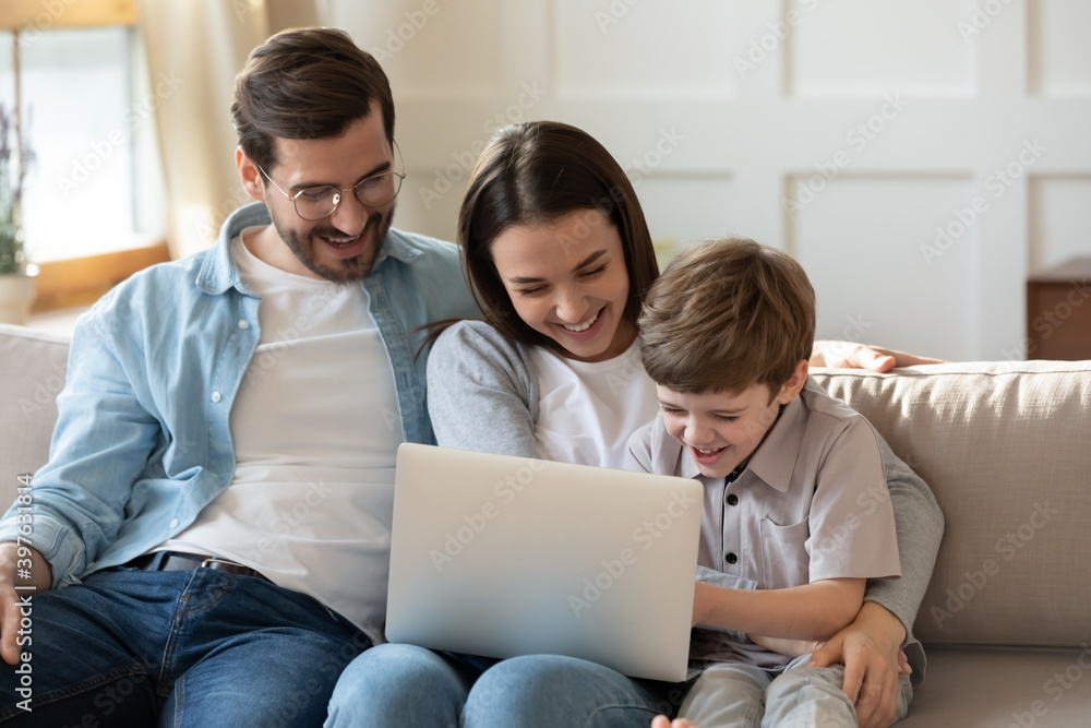 Overjoyed young parents with small son relax on sofa in living room laugh watching funny video on laptop. Smiling family with little boy child enjoy weekend at home, have fun use modern computer Overjoyed young parents with small son relax on sofa in living room laugh watching funny video on laptop. Smiling family with little boy child enjoy weekend at home, have fun use modern computer