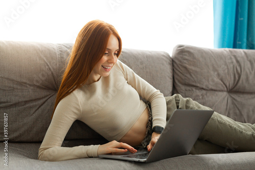 beautiful young female with red hair lies at home on the couch with a laptop