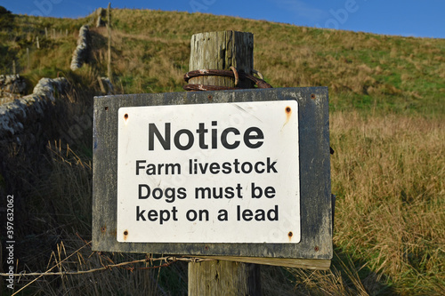 Isolated white sign with blurred background of stone wall, grass and blue sky. Text: Notice. Farm livestock. Dogs must be kept on a lead. Taken in Scotland, UK.