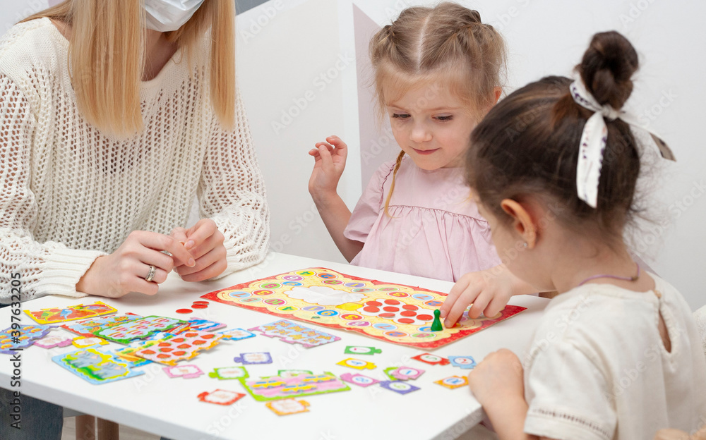 A kindergarten teacher plays a board game with two little girls. Mom plays with little daughters A kindergarten teacher plays a board game with two little girls. Mom plays with little daughters