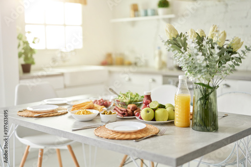 Design ideas. Close up of vase with flowers and Latin style breakfast on the kitchen table. Modern bright white kitchen interior with wooden and white details