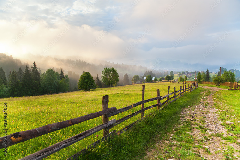 Morning mountain landscape, wooden fence along meadow and village road, mountains in fog. Ukraine, Verkhovyna.

