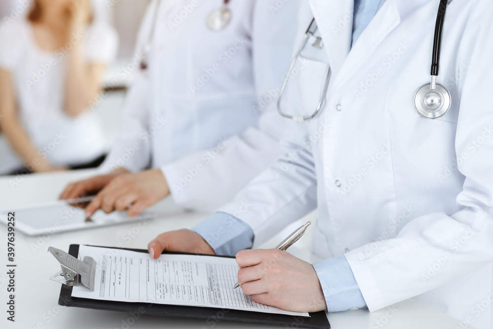 Unknown woman-doctors at work with patient at the background. Female physicians filling up medical documents or prescription while standing in sunny hospital reception desk, close-up. Health care Unknown woman-doctors at work with patient at the background. Female physicians filling up medical documents or prescription while standing in sunny hospital reception desk, close-up. Health care