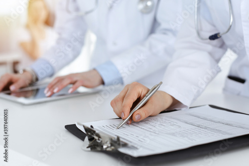 Unknown woman-doctors at work with patient at the background. Female physicians filling up medical documents or prescription while standing in hospital reception desk  close-up. Health care concept