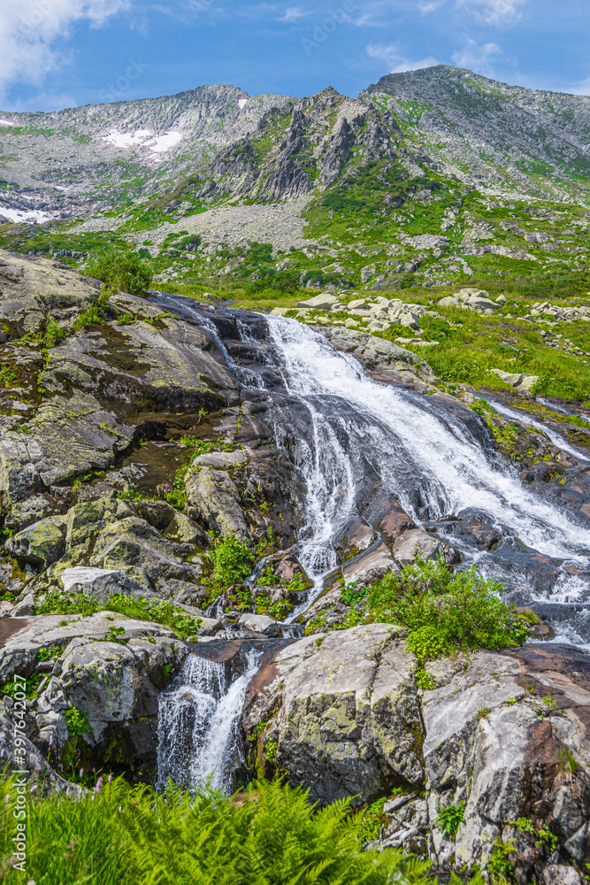 Rapid stream in mountain valley among grassy banks. Small waterfall in green meadow.