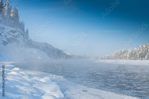 Frosty fog over winter river with snow and forest on bank. First ice on lake on cold day.
