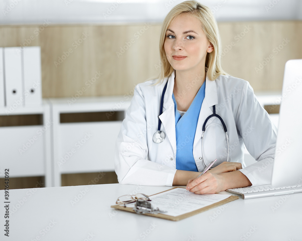 Portrate of woman-doctor at work while sitting at the desk in clinic. Blonde cheerful physician ready to help patients Portrate of woman-doctor at work while sitting at the desk in clinic. Blonde cheerful physician ready to help patients