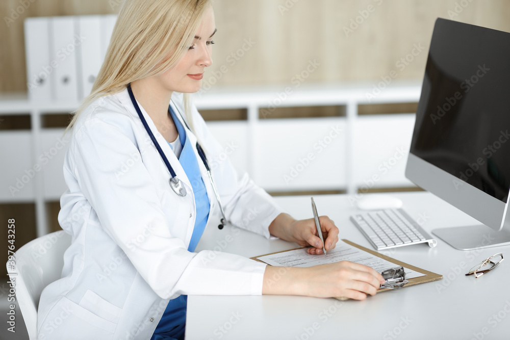Woman-doctor at work while sitting at the desk in hospital or clinic. Blonde cheerful physician filling up medication history record form Woman-doctor at work while sitting at the desk in hospital or clinic. Blonde cheerful physician filling up medication history record form