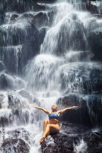 Travel in Bali jungle. Beautiful young woman sitting on rock under falling spring water, enjoy tropic cascade waterfall. Asian nature, day trip, walking adventure, fun on family summer vacation