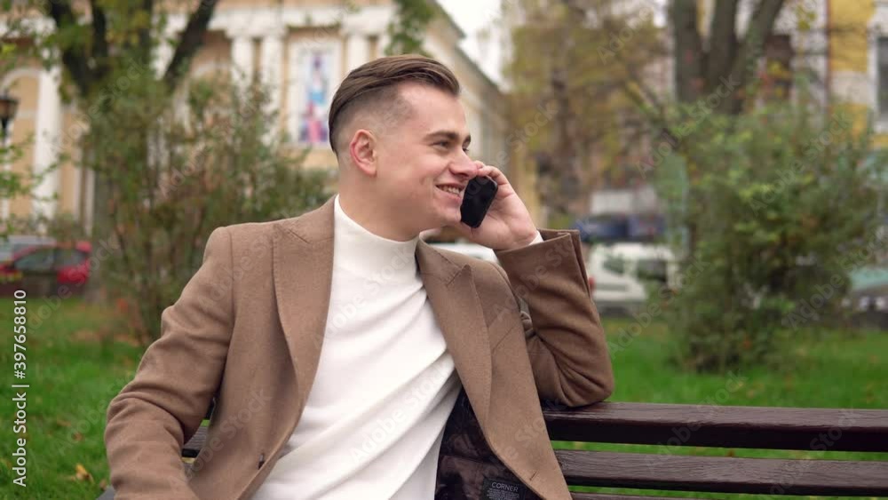 Elegant smiling man talking on the phone while sitting on a bench in the street
