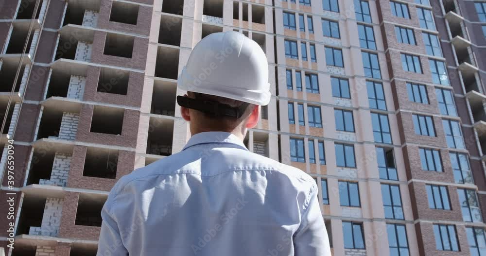 A man in a white protective helmet enters the frame and stops in front of the apartment building construction site. The camera shoots a person from the back. A man in a white protective helmet enters the frame and stops in front of the apartment building construction site. The camera shoots a person from the back.