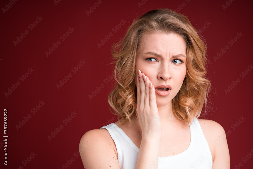 Blond woman with curly hair looks puzzled or having toothache in white tank top on red background. Blond woman with curly hair looks puzzled or having toothache in white tank top on red background.