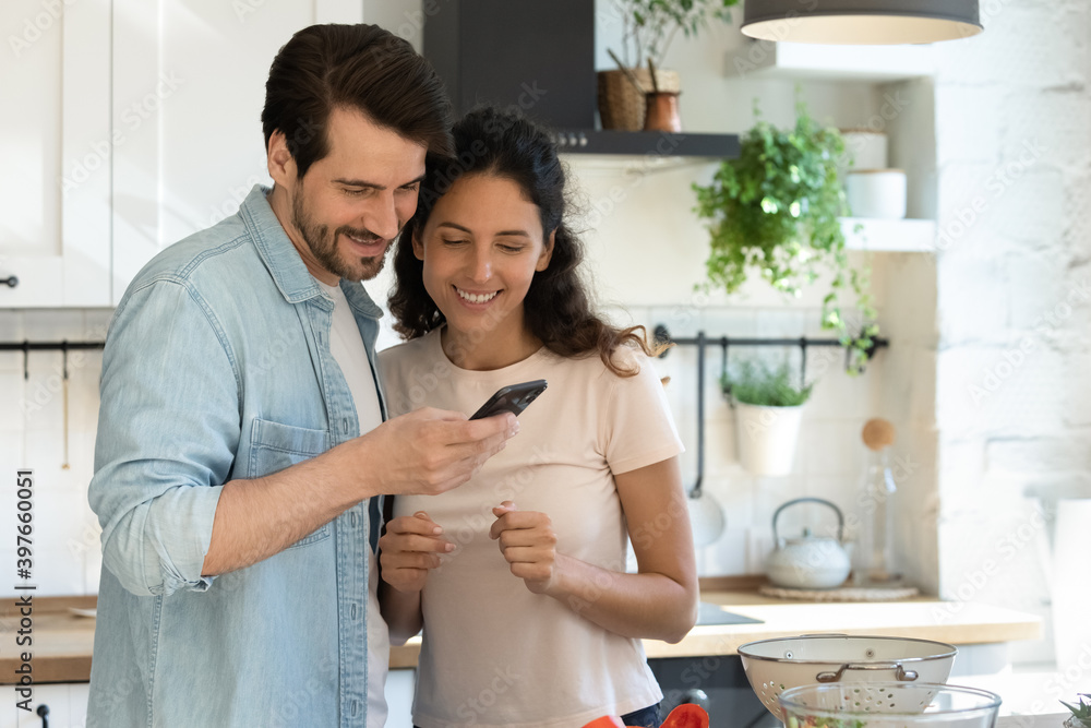 Close up happy couple using phone together, standing in kitchen at home, cooking, searching recipe in internet, browsing apps, ordering food online, smiling wife and husband enjoying leisure time Close up happy couple using phone together, standing in kitchen at home, cooking, searching recipe in internet, browsing apps, ordering food online, smiling wife and husband enjoying leisure time