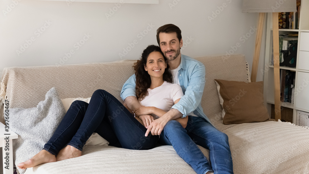 Portrait of young wife and husband hugging, sitting on couch, smiling attractive woman and man cuddling, looking at camera, posing for family photo at home, relaxing on sofa, enjoying lazy weekend