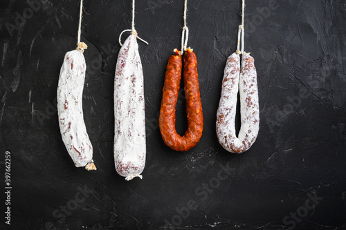 Spanish salami, longaniza and chorizo sausages hang from a rack on black textured background with copy space