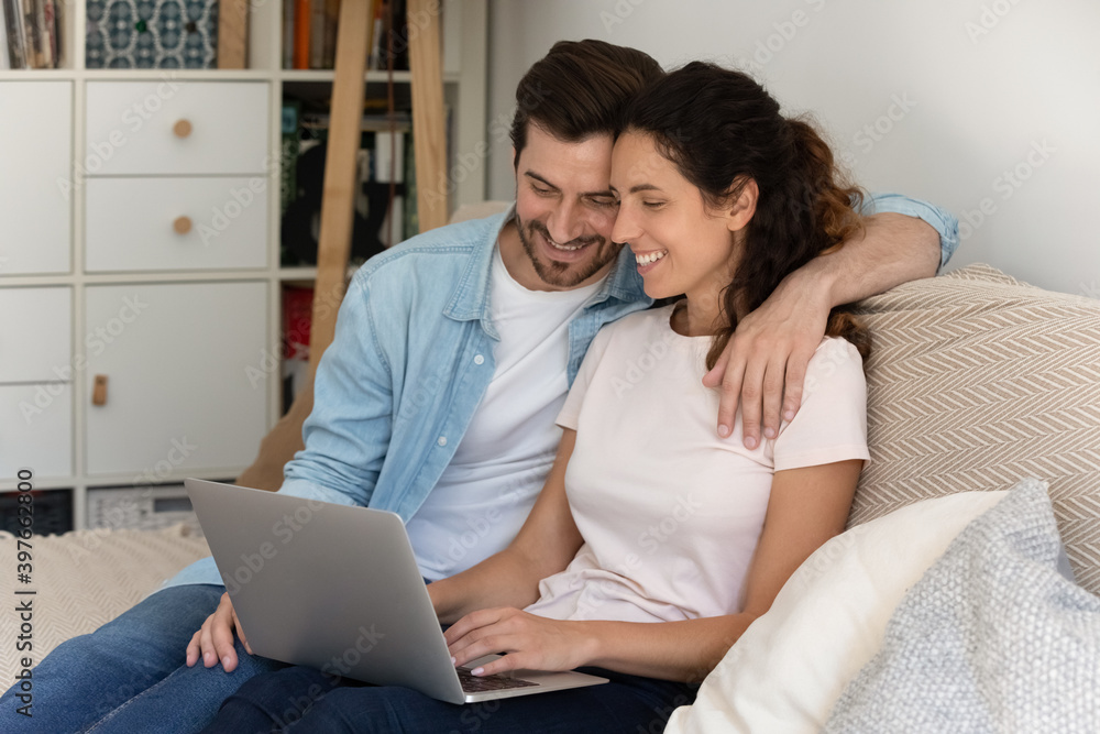 Happy wife and husband hugging, using laptop together at home, sitting relaxing on cozy couch, enjoying leisure time with device, smiling young woman and man browsing apps, shopping online