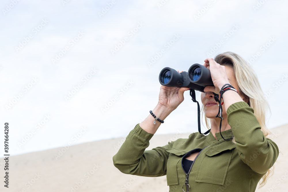 Woman looks through binoculars, standing in nature against a background of trees and sand.