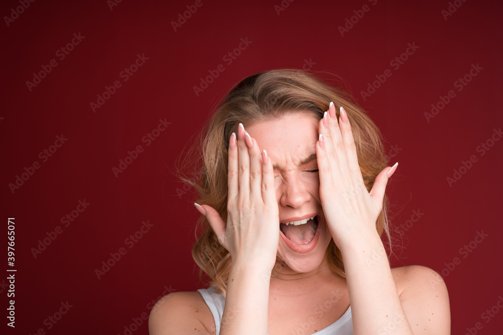 Blond woman screaming and covering her eyes with both hands in white tank top on red background. Frightened. Blond woman screaming and covering her eyes with both hands in white tank top on red background. Frightened.