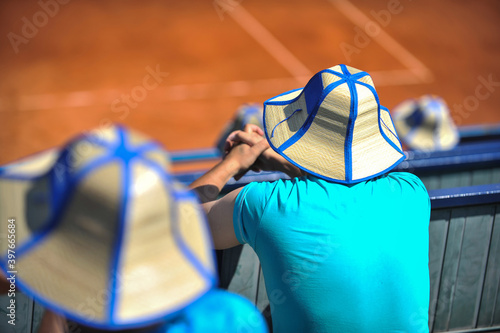Man wears a straw sun hat watching a tennis game during a sunny and hot summer day.