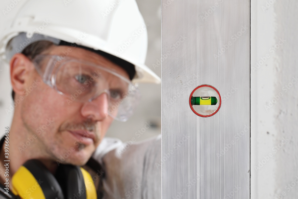 Φωτογραφία man worker looks at the spirit level checks the wall with hard hat, glasses and