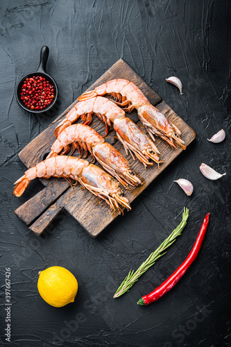 Fine selection of tiger prawns on cutting board with spices, pepper and lemon on black textured background, top view