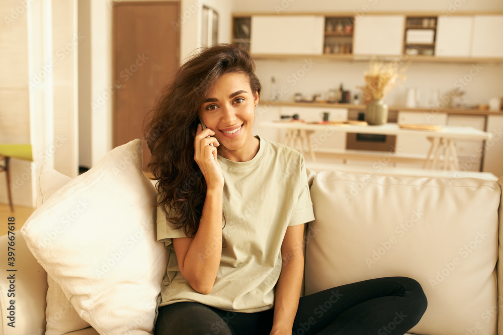 Indoor shot of adorable Latin teenage girl in casual clothing ordering food delivery, sitting on comfortable sofa with kitchen in background, speaking to operator on cell phone. Technology and gadgets Indoor shot of adorable Latin teenage girl in casual clothing ordering food delivery, sitting on comfortable sofa with kitchen in background, speaking to operator on cell phone. Technology and gadgets