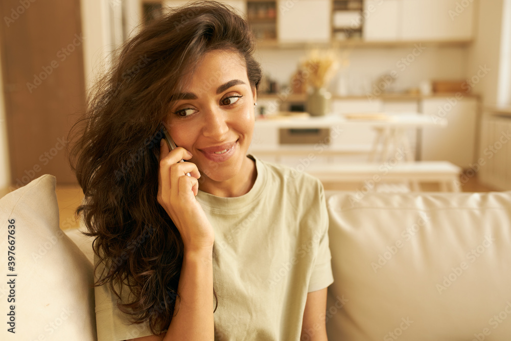 Close up image of beautiful young Arabic dark haired woman resting at home on weekend, sitting on couch with mobile at her ear, listening attentively and smiling, receiving good news over phone Close up image of beautiful young Arabic dark haired woman resting at home on weekend, sitting on couch with mobile at her ear, listening attentively and smiling, receiving good news over phone