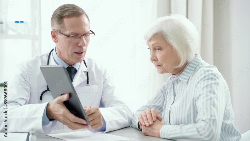 Medical help. Middle aged male doctor consulting elderly woman, holding tablet pc and prescribing treatment to his patient, sitting at the table in his office Medical help. Middle aged male doctor consulting elderly woman, holding tablet pc and prescribing treatment to his patient, sitting at the table in his office