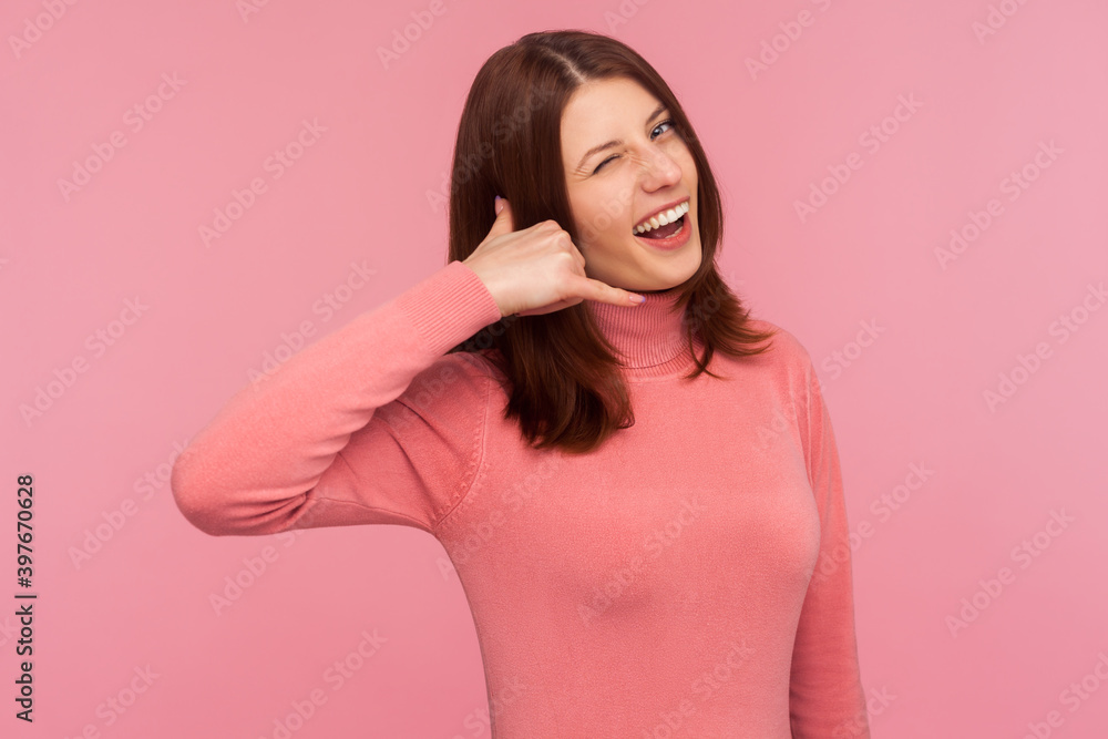 Cheerful playful woman with brown hair in pink sweater showing phone gesture and winking, asking to call her. Indoor studio shot isolated on pink background Cheerful playful woman with brown hair in pink sweater showing phone gesture and winking, asking to call her. Indoor studio shot isolated on pink background