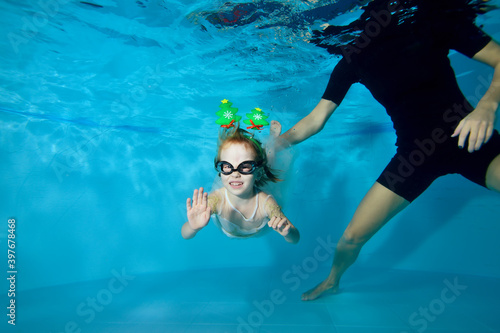 A little girl in a dress is swimming under the water in the pool. Christmas decorations on the head. Classes with the baby. Swimming lesson. Healthy lifestyle. Horizontal orientation