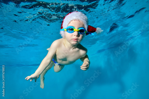 Funny boy in a Santa Claus hat and glasses in the pool underwater. The kid learns to have fun diving under water. Swimming lessons with a child. Healthy lifestyle. Horizontal orientation