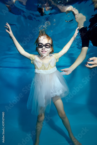 Portrait of a beautiful little girl underwater at the bottom of the pool. She smiles and looks at the camera. Baby learns to dive. Swimming lessons with a child. Bodily exercises. photo