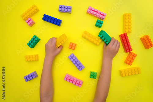Children's hands hold the details of the constructor on a yellow background. Multi-colored construction blocks.