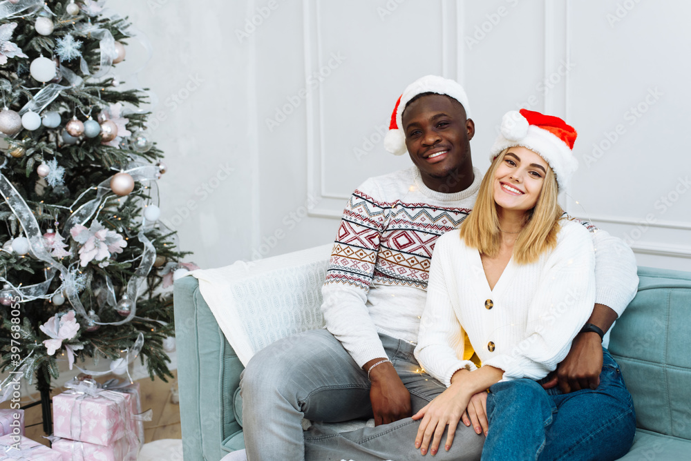 Fototapeta premium A young multiethnic couple in santa hats are sitting on a sofa in a bright interior next to a tree. Man and woman looking at the camera, smiling