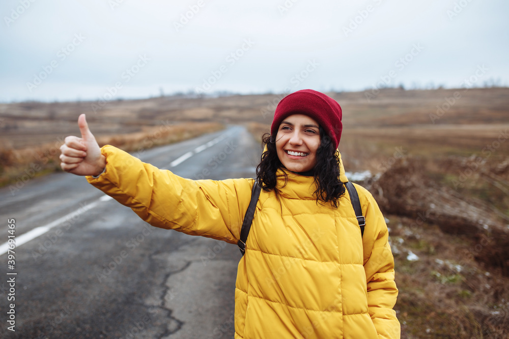 Female tourist with a backpack wearing yellow jacket and red hat catches a car on the road. Young woman travels during winter or late autumn season. Hitch-hiking, trip, thumb up, travelling concept.