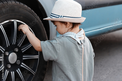 Stylish, happy child walks in the summer park by the river in a straw hat, inspects a car wheel