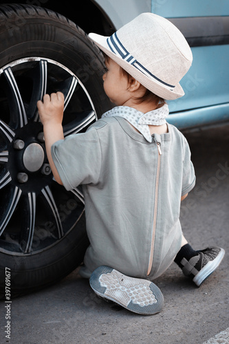 Stylish, happy child walks in the summer park by the river in a straw hat, inspects a car wheel