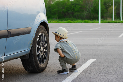 Stylish, happy child walks in the summer park by the river in a straw hat, inspects a car wheel