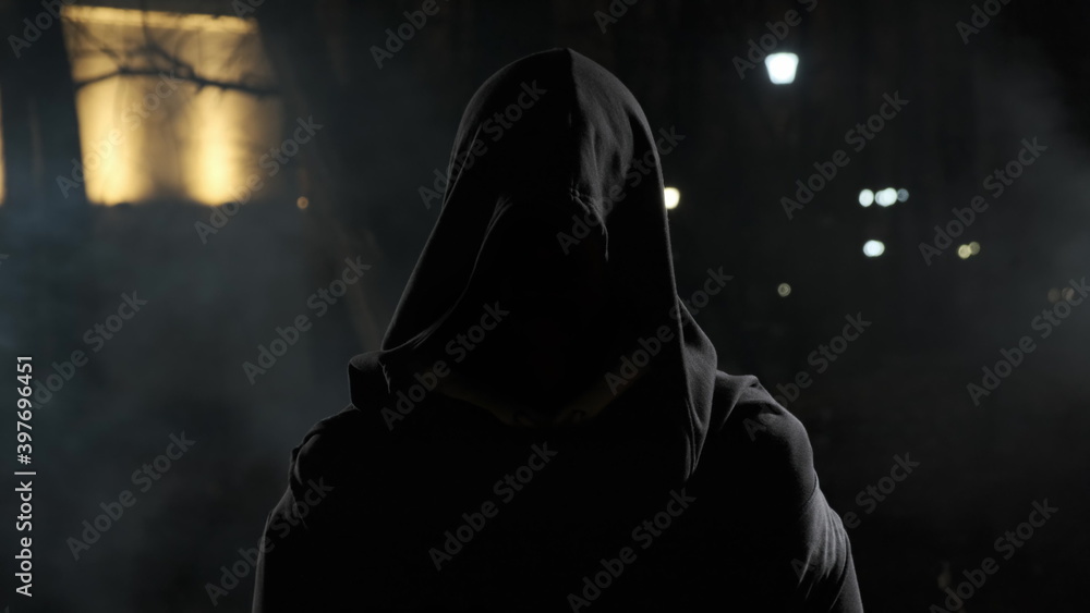 man in street at night in hood stands in front of camera. Scary ghost in night park with smoke around. Horror of the city with black hood. - obrazy, fototapety, plakaty 