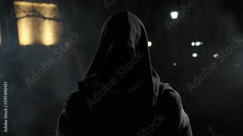 man in street at night in hood stands in front of camera. Scary ghost in night park with smoke around. Horror of the city with black hood. photo