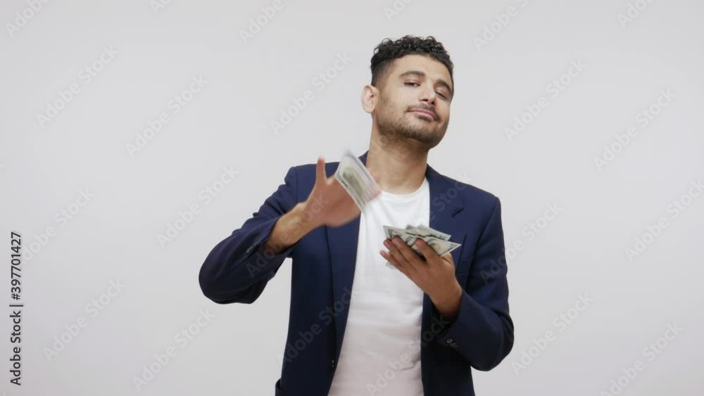 Rich mercantile bearded man in business suit scattering dollar bills, looking at camera with satisfied expression, bragging with big salary. Indoor studio shot isolated on gray background