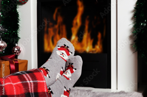 Girl warms her feet near the fireplace fire at home. Woman legs are covered with blanket and wear decorative Xmas socks. Winter and cold weather concept