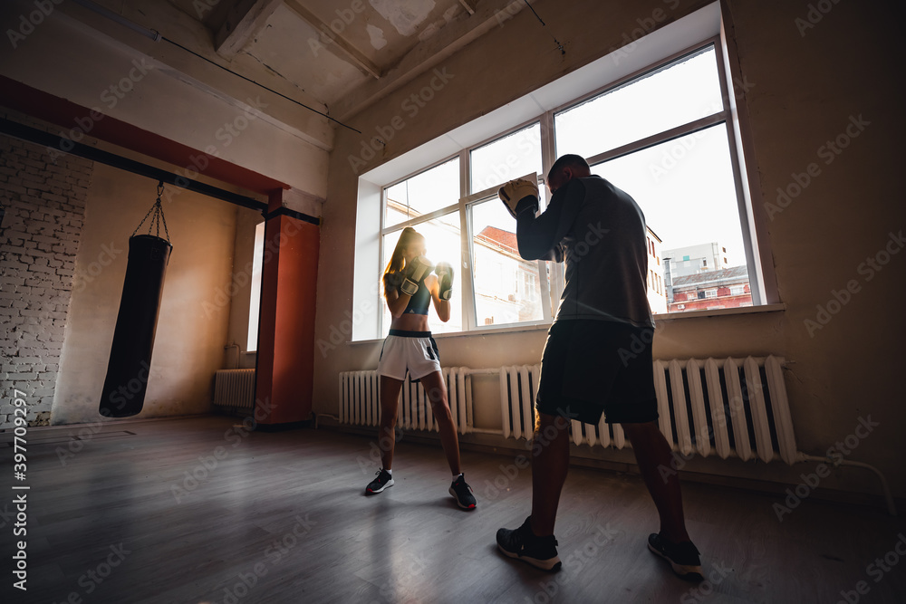 A man with boxing paws on his hands teaches the technique of hitting a novice girl in a light gym