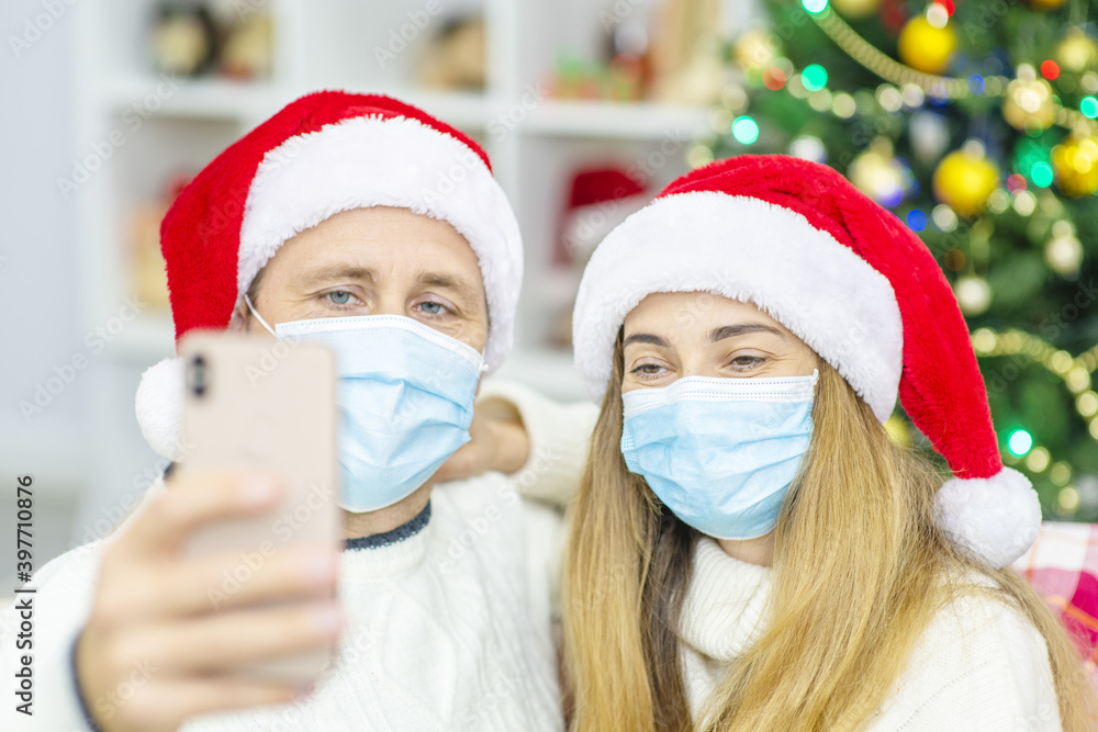 A married couple with medical masks are sitting on the sofa in Santa hats against the background of a Christmas tree and congratulating relatives with Christmas (New Year) online using a phone. A married couple with medical masks are sitting on the sofa in Santa hats against the background of a Christmas tree and congratulating relatives with Christmas (New Year) online using a phone.