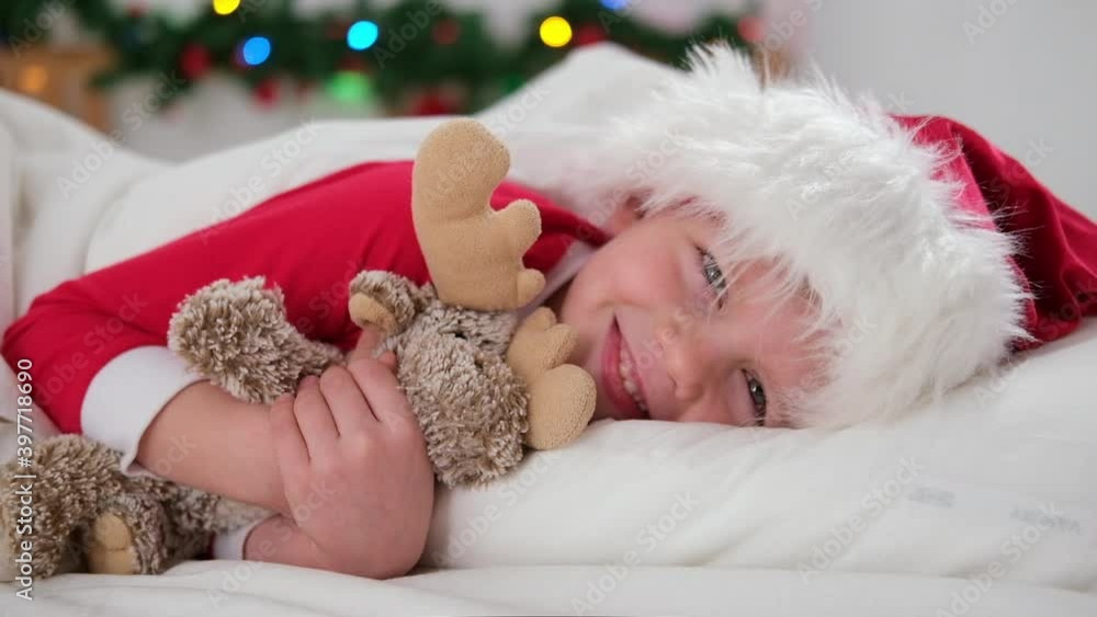 Child wakes up before Christmas. Little boy in Santa hat and Christmas pajamas lying in white bed hugging plush toy, opening eyes. New Year and Holidays. morning happiness Child wakes up before Christmas. Little boy in Santa hat and Christmas pajamas lying in white bed hugging plush toy, opening eyes. New Year and Holidays. morning happiness